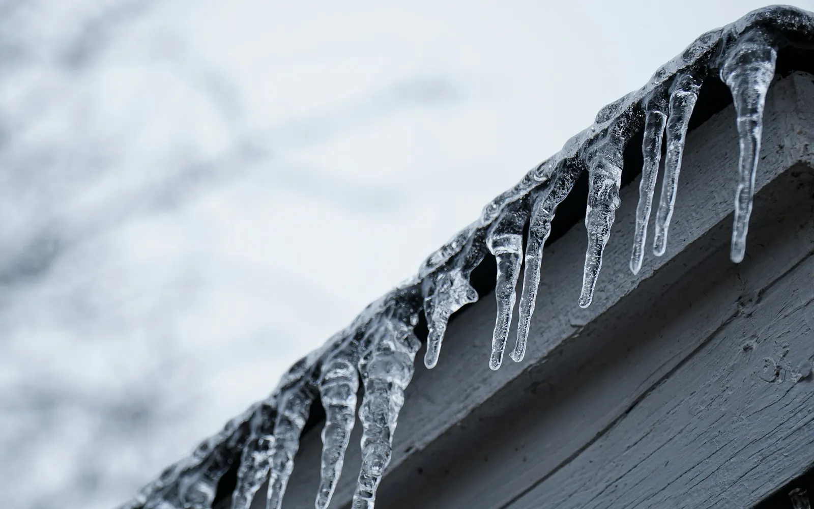 Long icicles hang from the snow-dusted edge of a roof eave, a sign of ice building up at the roofline.